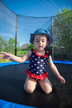 A cheerful little girl in a charming swimsuit is happily kneeling on a colorful trampoline, ready for fun and games