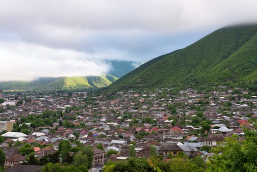 Shaki City in Azerbaijan sits nestled amidst rolling hills and abundant greenery, with clouds enveloping the landscape, creating a serene atmosphere.