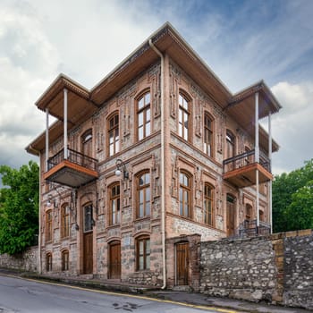 An elegant historic building stands in Shaki, Azerbaijan, featuring traditional architecture with detailed wooden balconies and large windows, against a backdrop of a cloudy sky.
