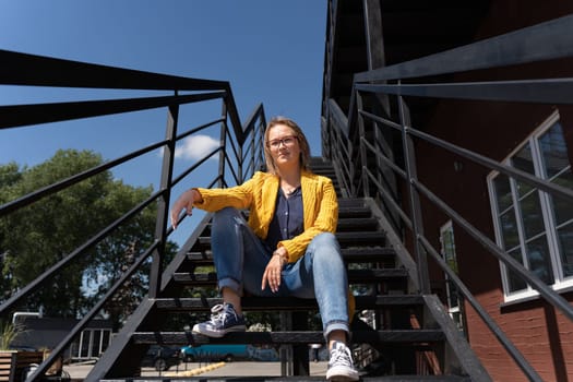 A woman in a bright yellow denim jacket relaxes on a set of metal stairs, with a view of the sky and a bridge in the background. She wears outdoor shoes and smiles, enjoying leisure time outdoors