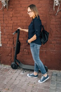 A woman in cool sneakers is standing on an electric scooter in front of a brick wall, wearing a denim blazer and a sleeveless top. Her waist is accessorized with a stylish belt