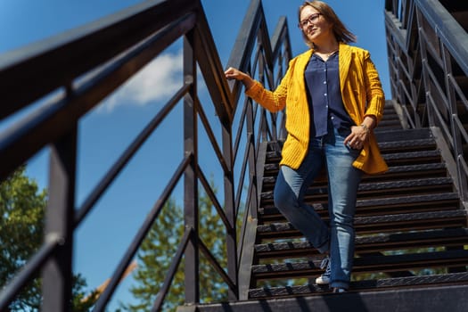 Smiling middle-aged woman in glasses descending stairs outdoors.