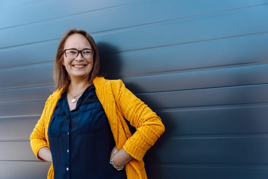 A happy woman with glasses, a yellow jacket and stylish hairstyle stands in front of a blue wall, flashing a smile and showcasing her vision care eyewear