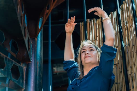 Woman in blue shirt raising arms against a bamboo fence background, natural light, industrial setting, concept of freedom
