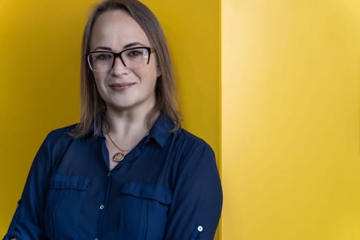 smiling 40-year-old woman in glasses wearing blue shirt on yellow background.