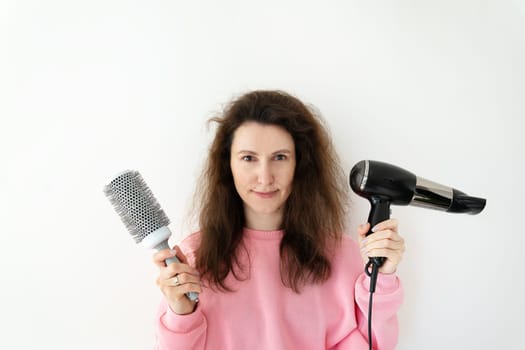 The girl holds a hair dryer and a comb in her hands, preparing for hair styling at home.Drying brown hair with a round brush for styling unruly hair