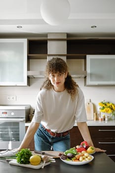 Curly-haired woman leaning against a kitchen table with fresh organic vegetables on it