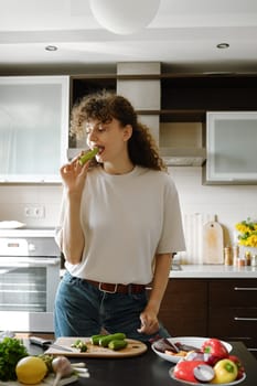 While preparing healthy and wholesome food, a woman bites fresh zucchini while standing in the kitchen