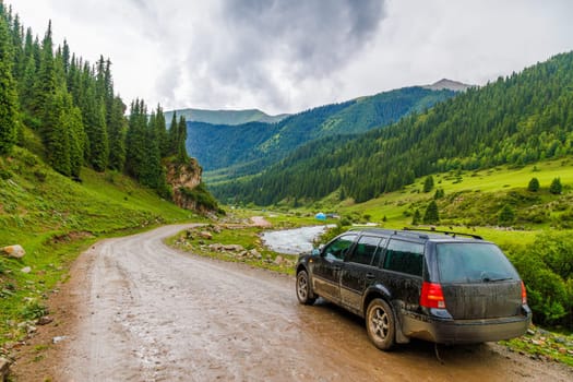 old black German station wagon car on a side of wet dirt road with green grass and forest covered mountain slopes at rainy summer day