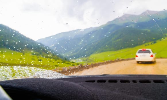 white car driving down the dirt road at rainy summer day in mountains, selective focus view from drivers perspective.