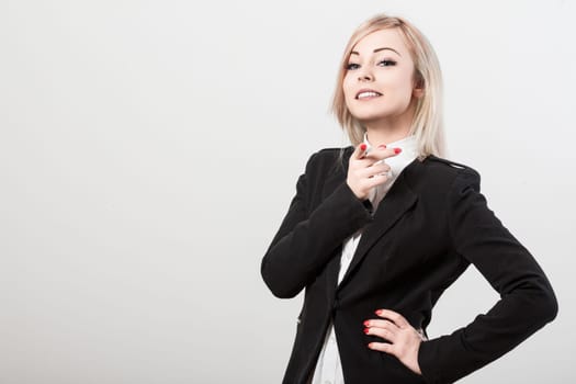Young businesswoman is posing with her hand on her hip and pointing with her index finger. She is wearing a black jacket and a white shirt