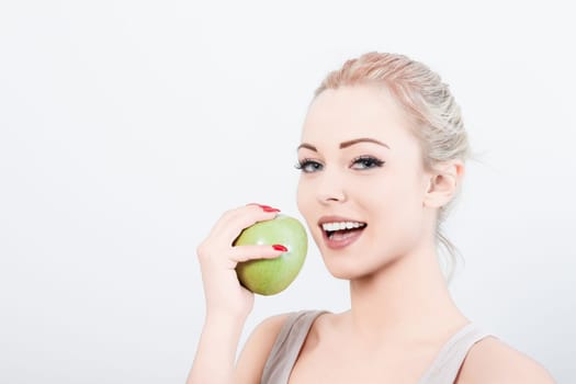 Cheerful woman with blonde hair tied back bites a fresh green apple against a white background, showcasing a healthy lifestyle with a bright smile