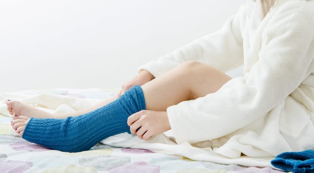 A person is sitting on a bed with a blue sock on their leg. The person is wearing a white robe and he is in a relaxed state