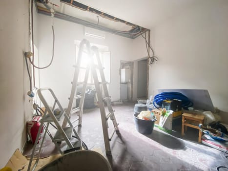 Interior of a room under renovation with exposed wiring, construction tools, debris, and damaged walls, showing ongoing reconstruction work.
