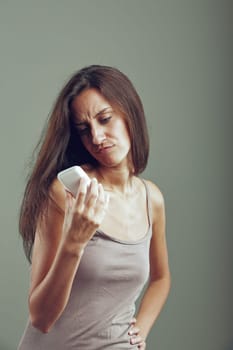 Worried young woman is frowning while reading a message on her smartphone. She is standing in front of a gray background
