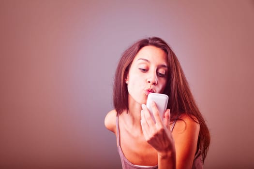 Young woman playfully sends a kiss through her smartphone screen against a pink backdrop, perfect for concepts like online dating and modern love