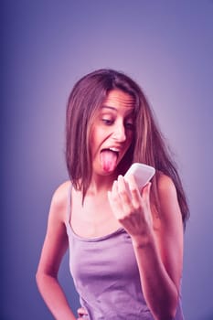 Young woman in her 20s playfully reacts to a text message on her smartphone, sticking out her tongue and looking disgusted against a purple background