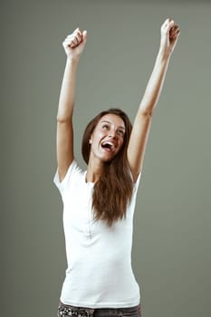 Young woman celebrating a win with arms raised in joy, radiating happiness and excitement in a grey studio setting