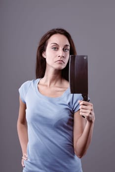 Serious young woman is holding a meat cleaver in her hand and posing for a portrait in a studio against a gray background