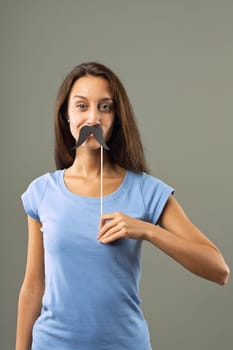 Young woman is playfully holding a fake mustache on a stick in front of her face, expressing a sense of lightheartedness and fun