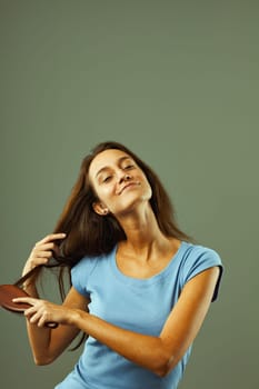 Young woman with long, dark hair happily brushes her hair with a wooden hairbrush, eyes closed, smiling serenely, showcasing her healthy hair care routine against a green background