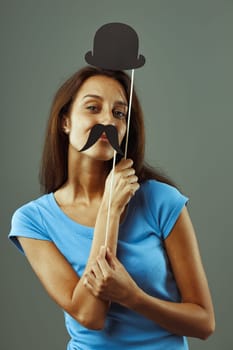 Young woman poses playfully with a fake mustache and bowler hat, radiating joy and happiness against a grey background