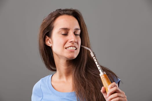 Young woman with long brown hair is holding a soldering iron close to her cheek with her eyes closed and teeth clenched