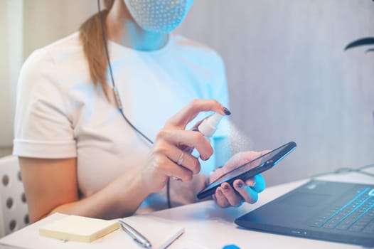 Woman disinfects the surface of the phone by sanitizer spray on the working place. Coronavirus concept. Woman in quarantine for coronavirus covid-19 working from home.