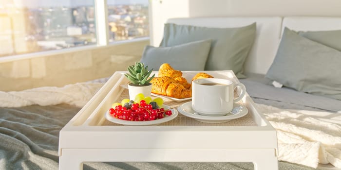 White Tray with tasty breakfast on bed in light bedroom with big windows in skyscraper. white table with tasty breakfast in bed at bed room in skyscraper