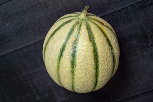 A large, round cantaloupe sits on a dark surface. The cantaloupe is green and yellow, with a green stem protruding from the top. Concept of freshness and natural beauty