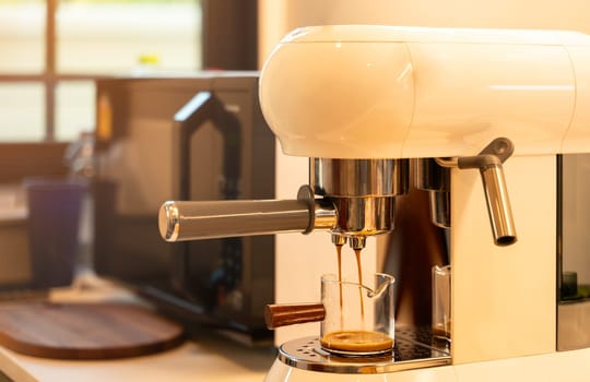 Espresso machine brewing aromatic coffee. Golden streams flowing into glass cup with rich crema in morning sunlight kitchen at home