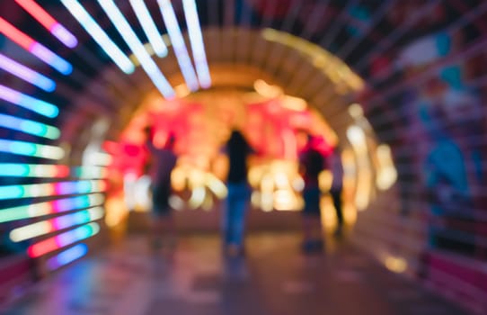Blurred abstract image of people gathered in vibrant neon lit tunnel. Radiating lines of blue, green, and purple lights frame background.