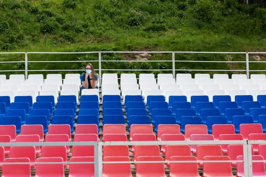 Lonely woman with protective mask on the empty stadium outdoor. Empty tribune due to pandemic Covid-19. Concept of pandemic life , empty stadiums, distance from viewers, safety.