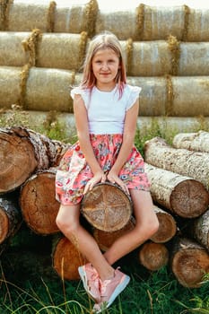 A young girl with long loose hair is sitting on a log in a pink dress with her legs dangling outside on a warm summer day