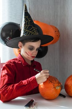 Happy teen boy in costume preparing for the Halloween celebration drawing a pumpkin. Halloween carnival or masquerade concept