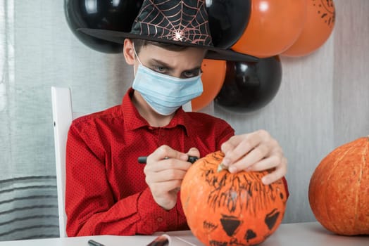 Teen boy in costume and face masks to protect against COVID-19 drawing a pumpkin for the Halloween celebration. Halloween carnival with new reality with pandemic concept.