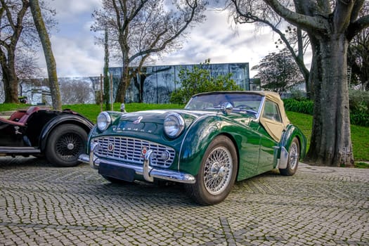 Lisbon, Portugal - Jan 20, 2024: A classic green Triumph TR3A soft top drophead car cobblestone driveways by another vintage vehicle on sunny spring day.