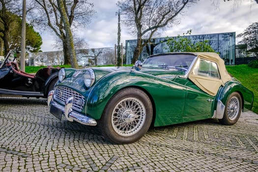 Lisbon, Portugal - Jan 20, 2024: Classic green Triumph TR3A soft top drophead car cobblestone driveways by another vintage vehicle on sunny spring day.