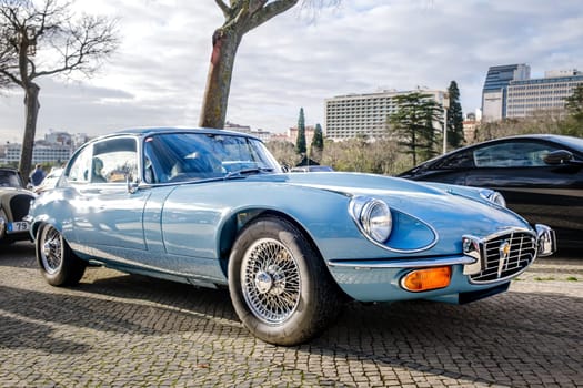 Lisbon, Portugal - Jan 20, 2024: A classic light-blue E-Type Jaguar Series 3 coupe car with chrome detailing parked on a cobblestone path near green park.