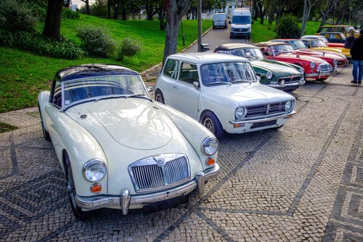 Lisbon, Portugal - Jan 20, 2024: A row of vintage cars of MG and Mini Cooper brand and others parked on cobblestone driveway near green park. MGA, 1973 Morris Mini Clubman, MGC Roadster, MGA TWIN CAM and others.