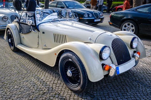 Lisbon, Portugal - Jan 20, 2024: A classic white or ivory Morgan Plus 4 sports car with chrome detailing parked on a cobblestone path near green park.