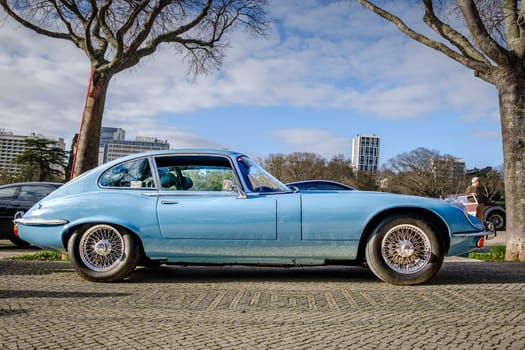 Lisbon, Portugal - Jan 20, 2024: Full lateral view of classic light-blue E-Type Jaguar Series 3 coupe car with chrome detailing parked on a cobblestone path near green park.
