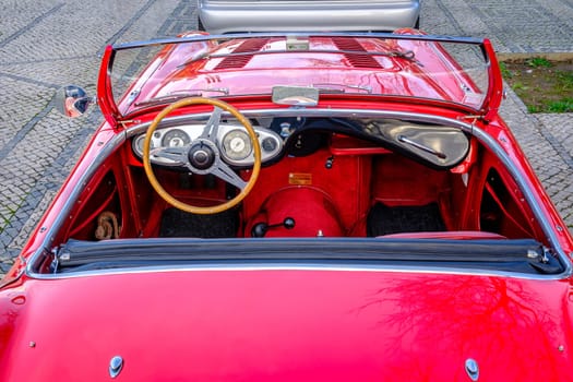 Lisbon, Portugal - Jan 20, 2024: Original red interior of red Austin-Healey 100 vintage convertible sports car parked on cobblestone pavement road. Red velvet and leather, wooden steering wheel, fold-down windshield