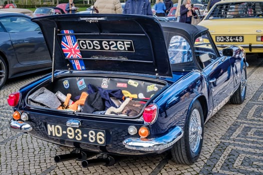 Lisbon, Portugal - Jan 20, 2024: Open trunk full of assorted items and stickers and rear side of classic Triumph Spitfire MK3 car parked on cobblestone path by another vintage vehicle showcasing the elegance and design of early automotive engineering