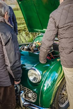 Three unrecognizable male adults stand by and look into open bonnet of vintage green car. Hood is lifted an engine compartment visible. Green grass in background