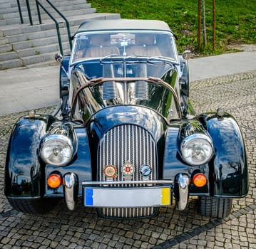 Lisbon, Portugal - Jan 20, 2024: A classic dark green Morgan Plus 4 sports car with chrome detailing parked on a cobblestone path near green park.