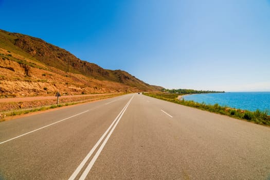 asphalt interstate road between yellow sedimentary rock mountain and large lake at sunny summer day, wide angle view