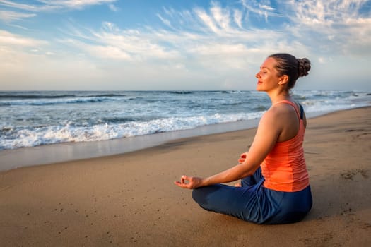 Young sporty fit woman doing yoga - meditating and relaxing in Padmasana Lotus Pose) with chin mudra outdoors at tropical beach on sunset