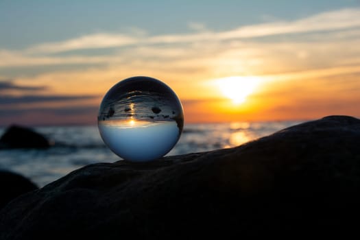 Glass ball on a rock at sunset on the beach, the sea and the setting sun are reflected in the ball