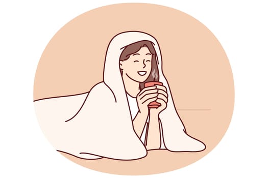 Girl with cup of hot coffee lies wrapped in blanket and smiles enjoying winter morning. Woman drinking hot drink with blanket to keep warm after walking on cold street for autumn mood concept